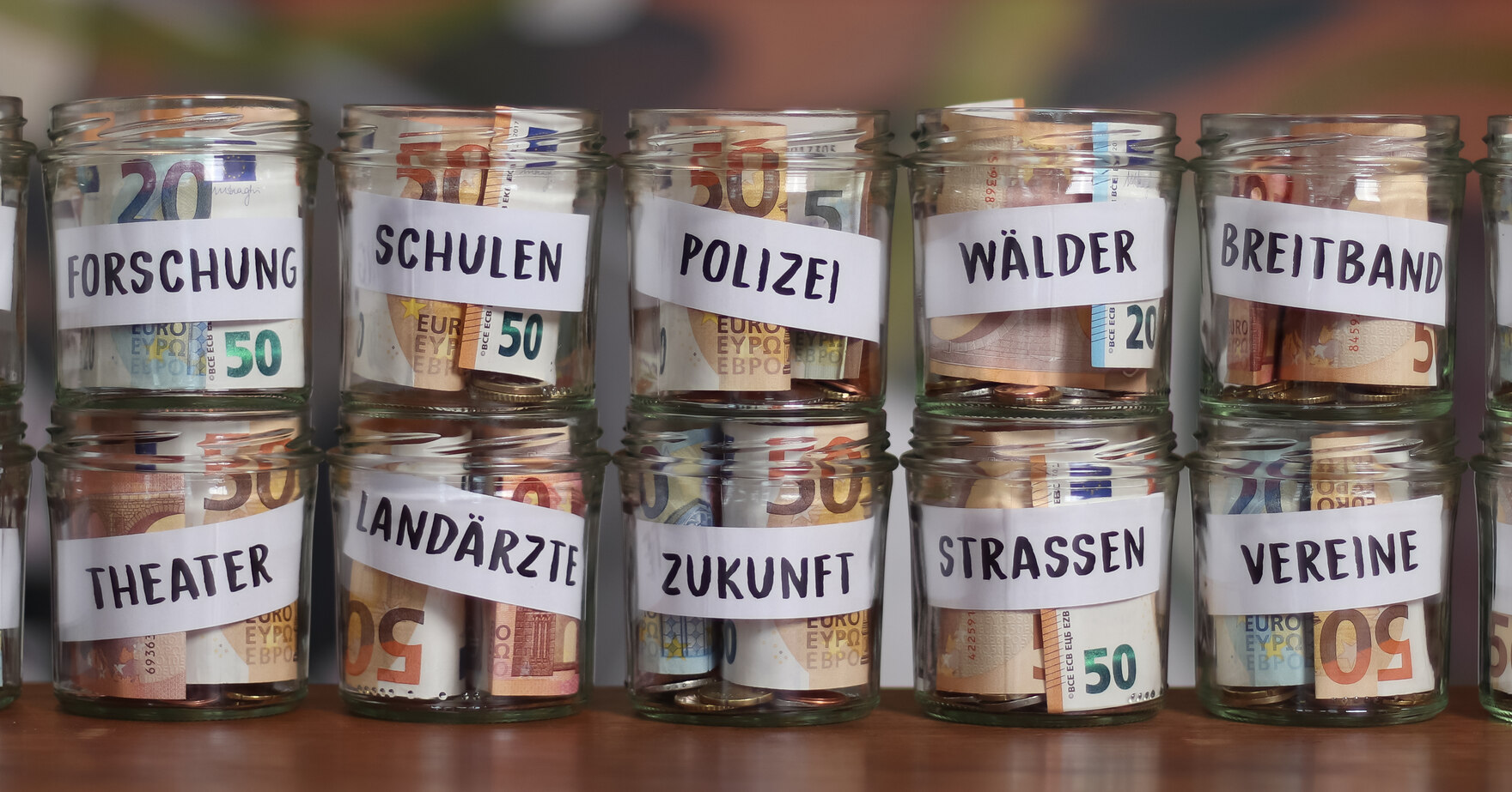 Gestapelte Gläser, in denen Geldscheine sind. Jedes Glas steht dabei für eine Verwendung der Gelder. Zum Beispiel Forschung, Schulen, Polizei und Kultur.